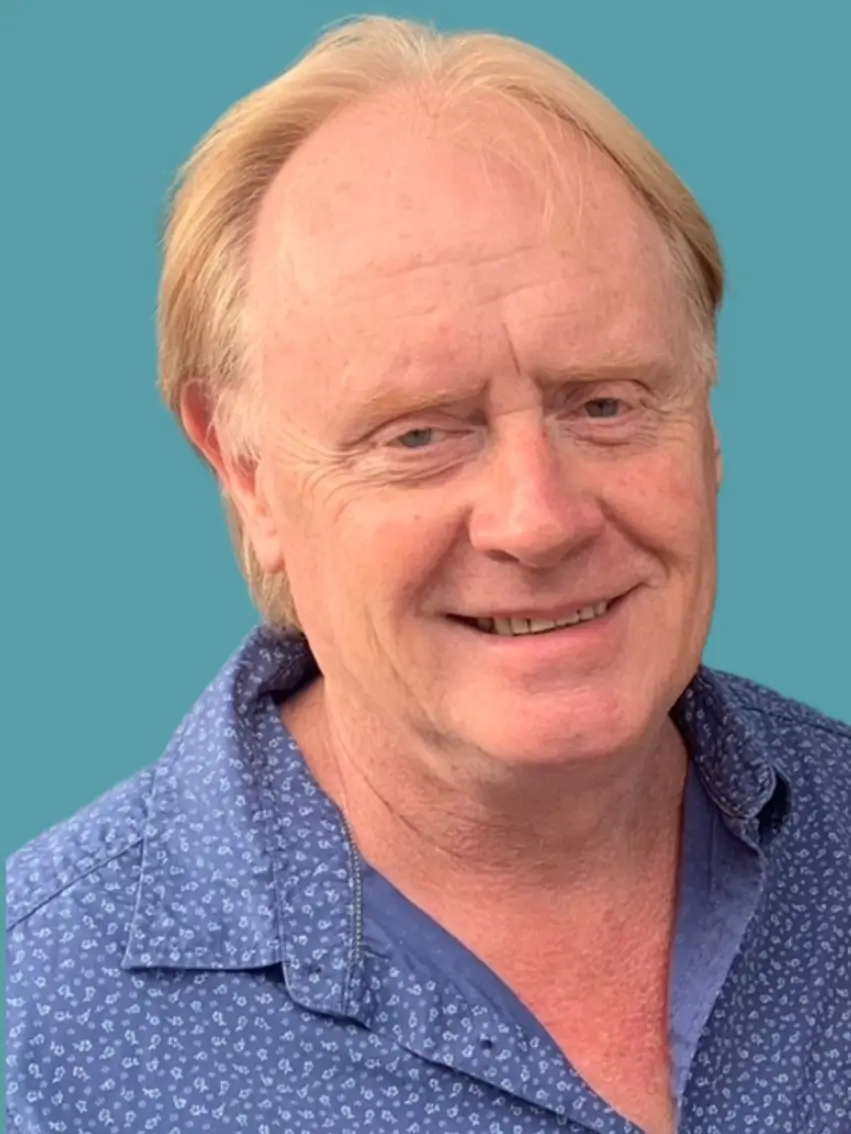 A middle-aged man with light skin and thinning blond hair smiles at the camera, wearing a blue patterned shirt against a solid teal background.
