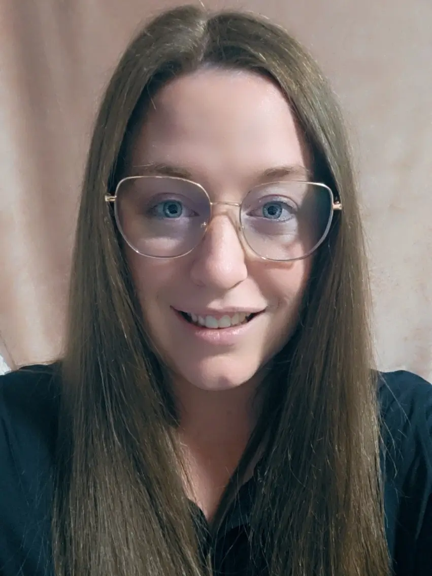 A person with long brown hair and glasses is smiling at the camera, wearing a dark top against a neutral background.
