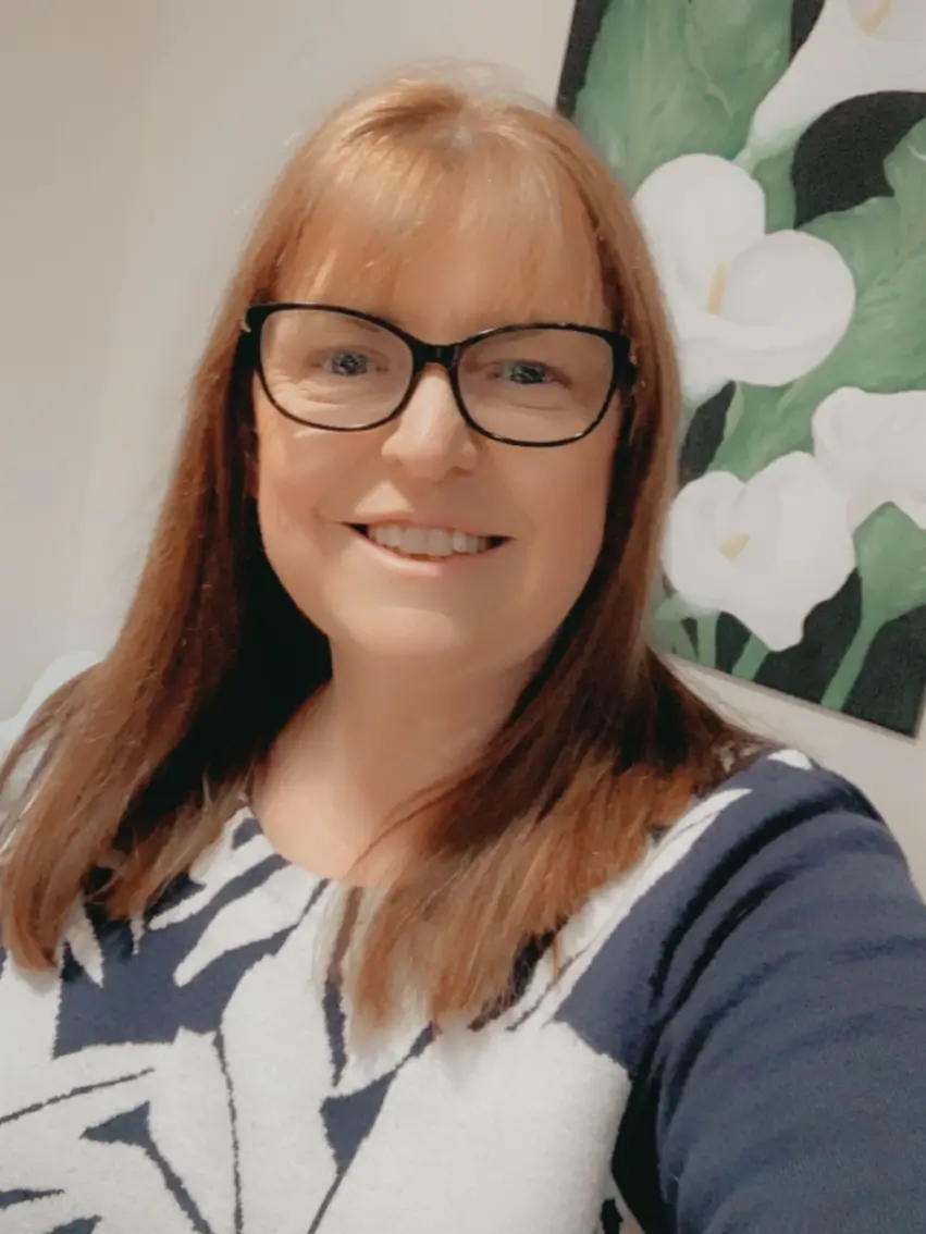 A woman with straight red hair and glasses smiles at the camera. She is wearing a patterned sweater, and there is a floral painting in the background.
