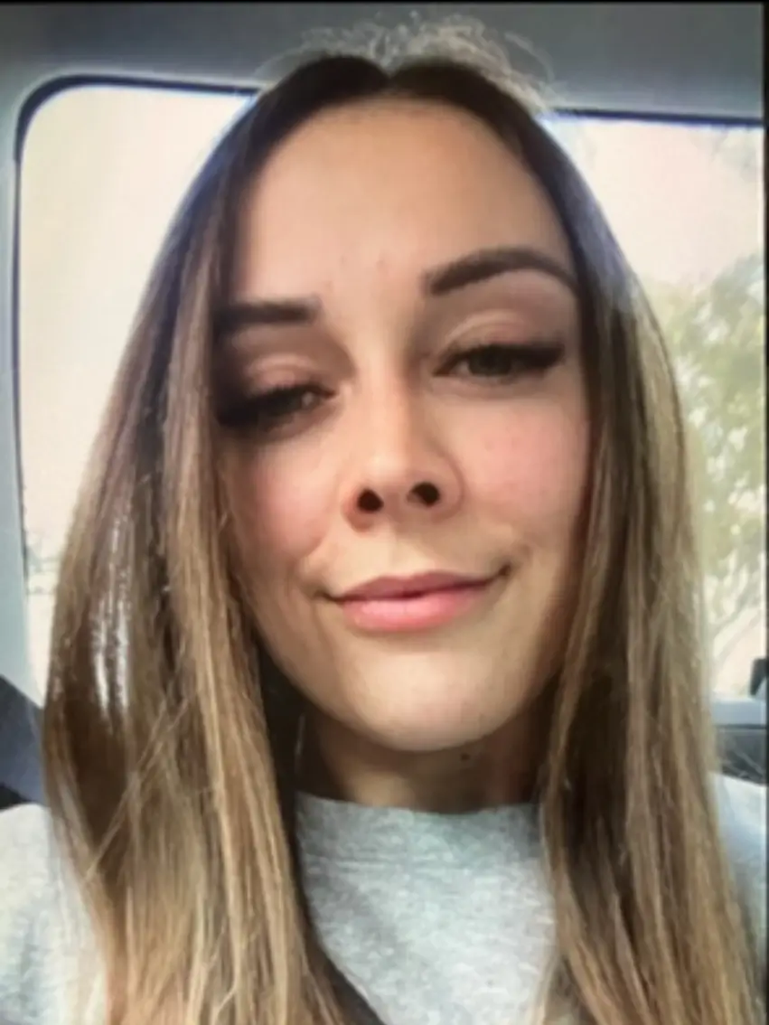 A woman with straight brown hair and a grey top sits in a car, facing the camera and smiling slightly.