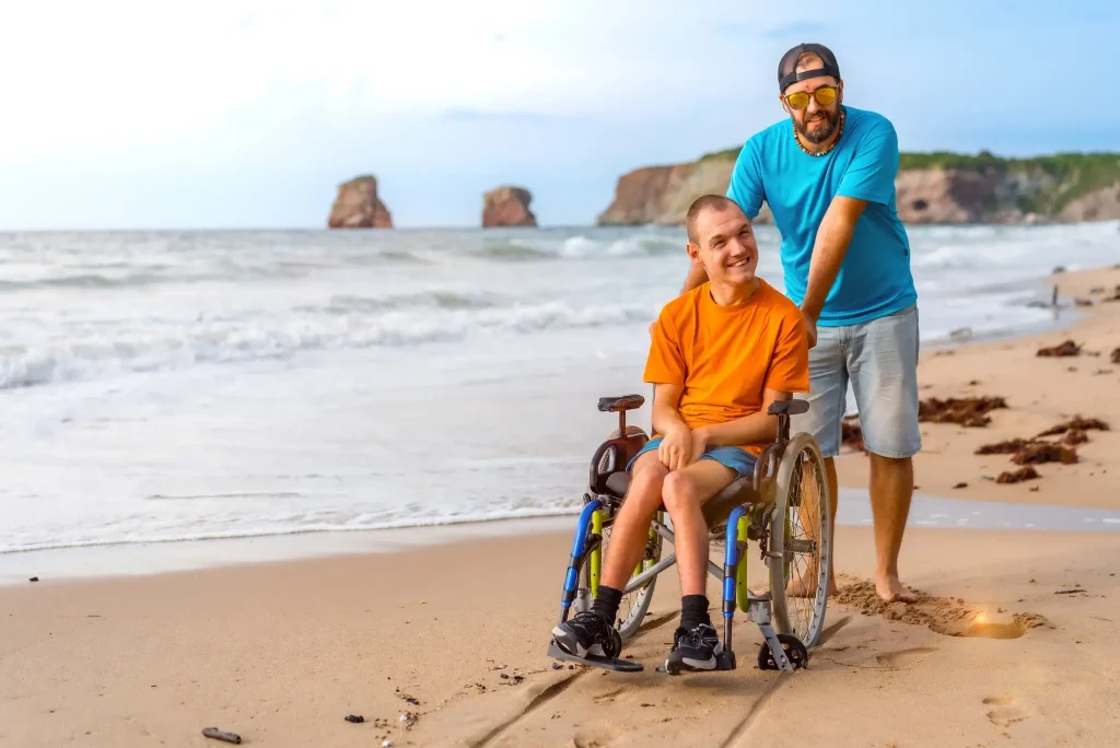 A man pushes a young man in a wheelchair along a sandy beach with waves and rock formations in the background.