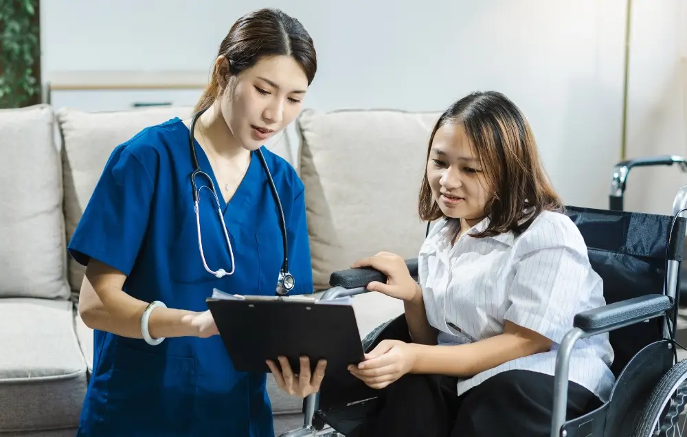 A nurse in blue scrubs shows a clipboard to a woman sitting in a wheelchair, both appearing focused on the document.