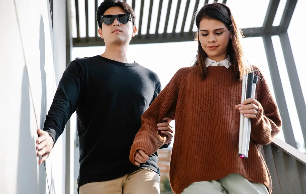 A man wearing sunglasses holds a woman's arm as they walk down stairs. The woman carries a rolled-up document in a tube.