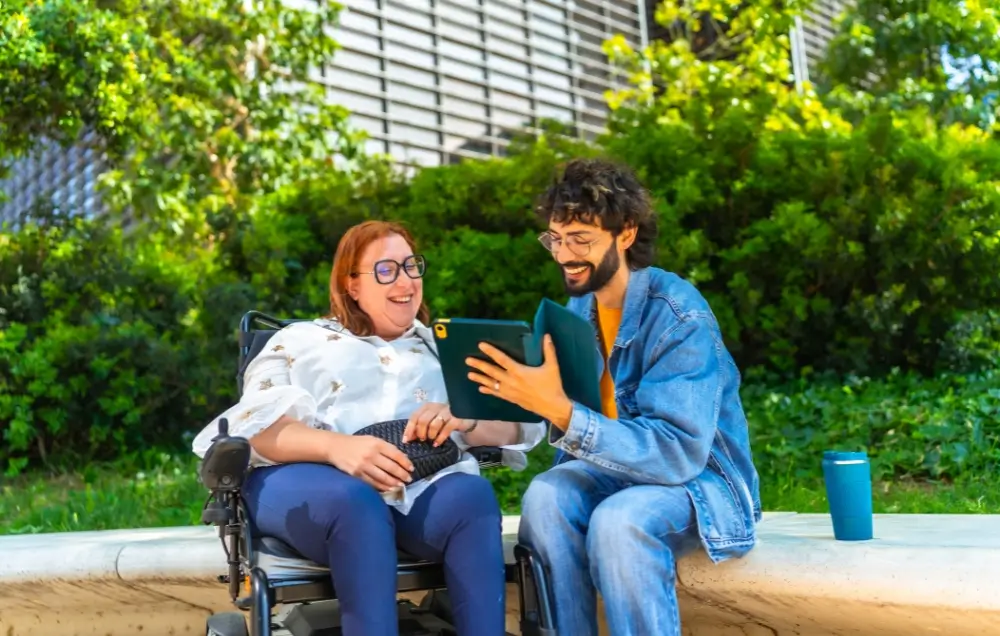 A woman in a wheelchair and a man sit on a bench outdoors, looking at a tablet together and smiling. A reusable cup is placed next to them.