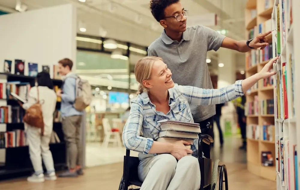 A woman in a wheelchair holding books reaches for a shelf in a library or bookstore while a man stands beside her, assisting. Other people browse in the background.