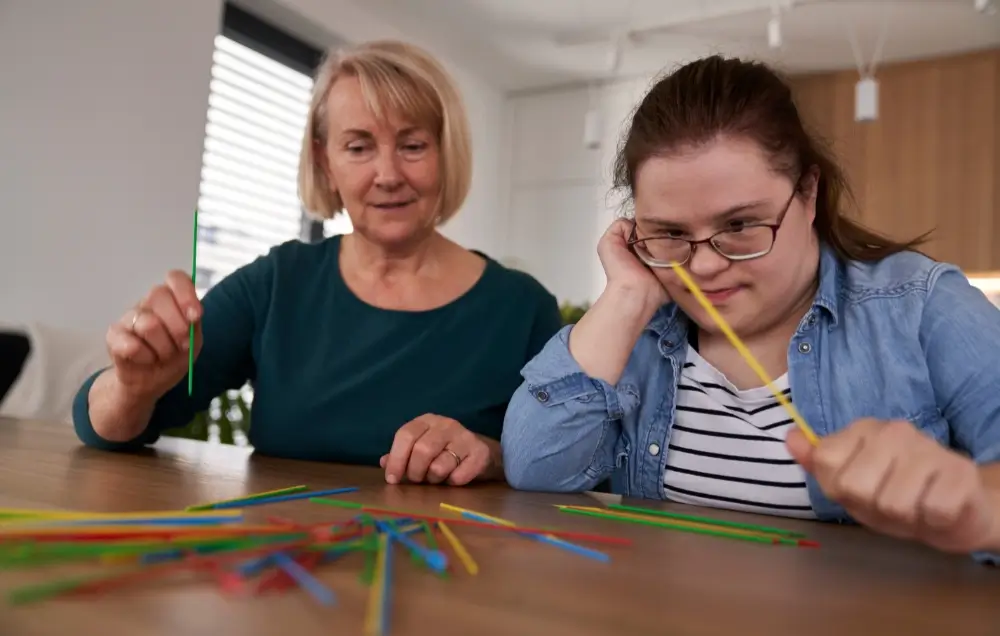 Two women sit at a table playing pick-up sticks; one observes while the other concentrates on picking up a stick.