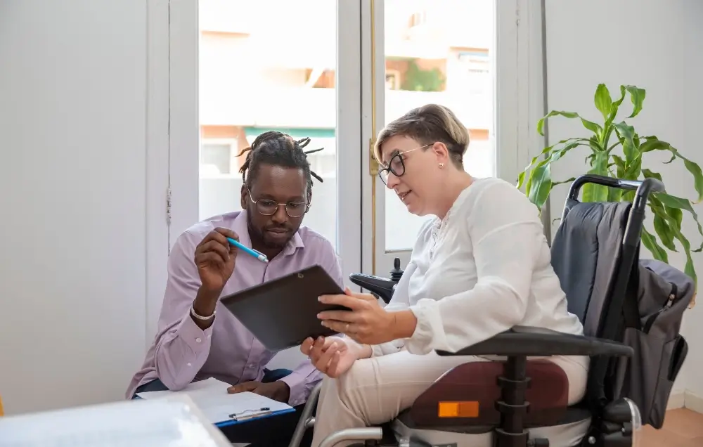 A man and a woman in a wheelchair sit together in an office, looking at a tablet and discussing documents.