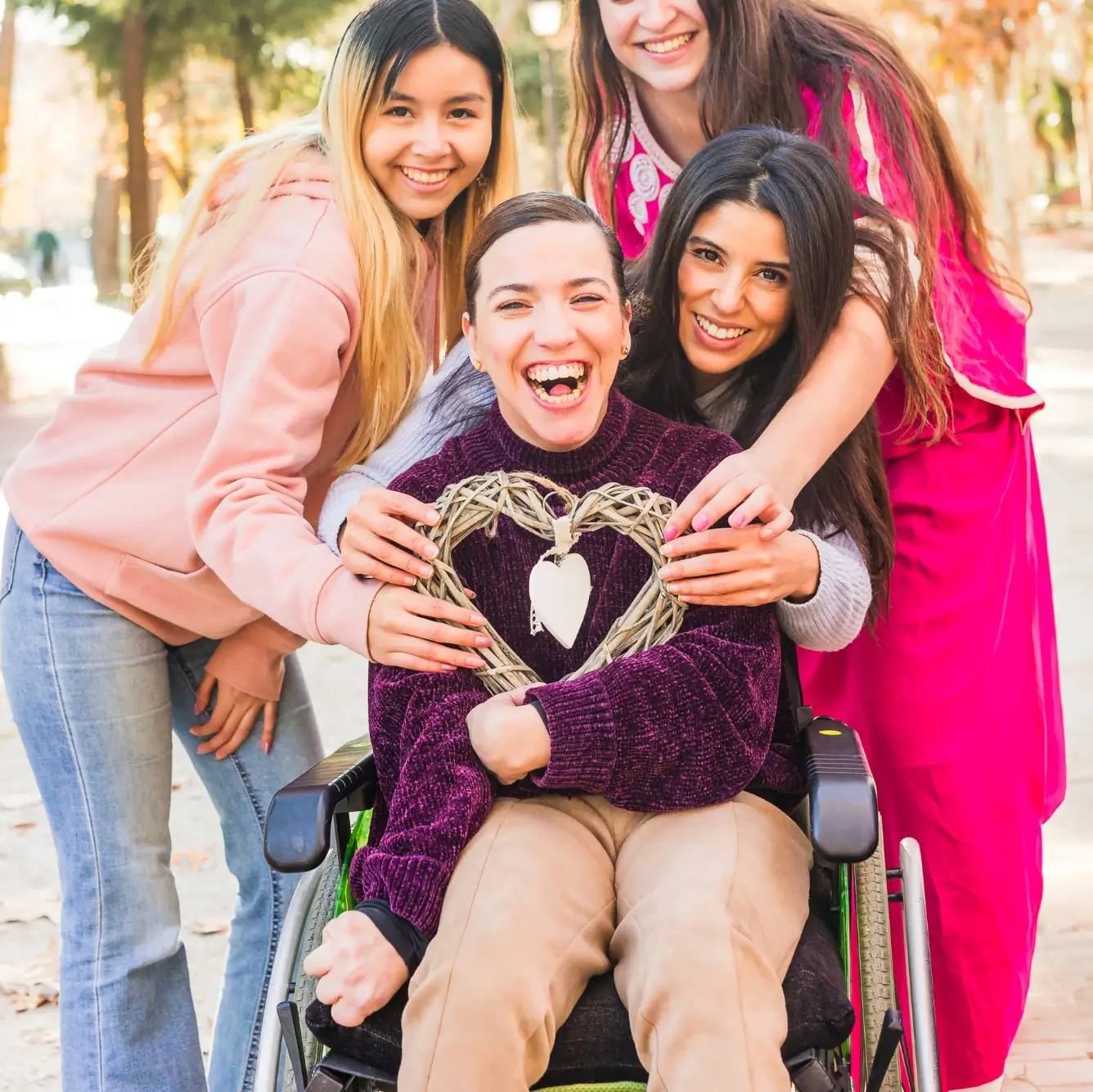 Four young women, one seated in a wheelchair, pose together outdoors. The woman in the wheelchair holds a decorative heart-shaped ornament. All are smiling at the camera.