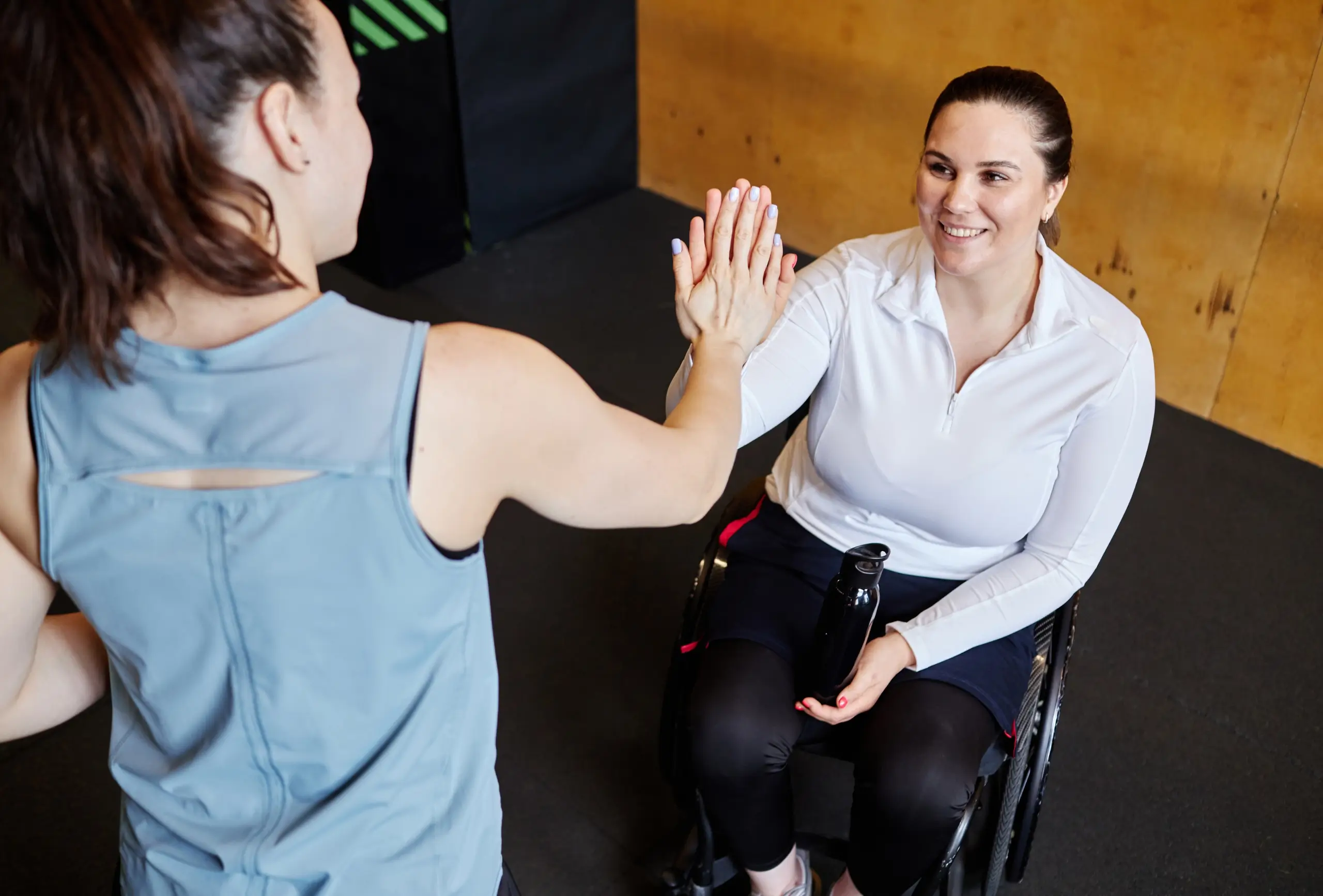 Two women are giving each other a high five; one is seated in a wheelchair with a water bottle, while the other stands facing her in a gym setting, highlighting the positive impact of choosing an NDIS provider and experienced nurses.