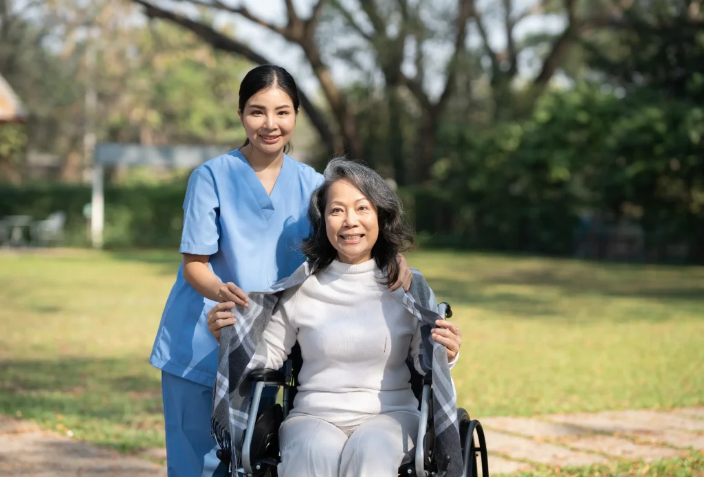 An experienced nurse in blue scrubs, representing an NDIS provider, stands behind an older woman in a wheelchair, gently placing a blanket around her shoulders in an outdoor park setting.