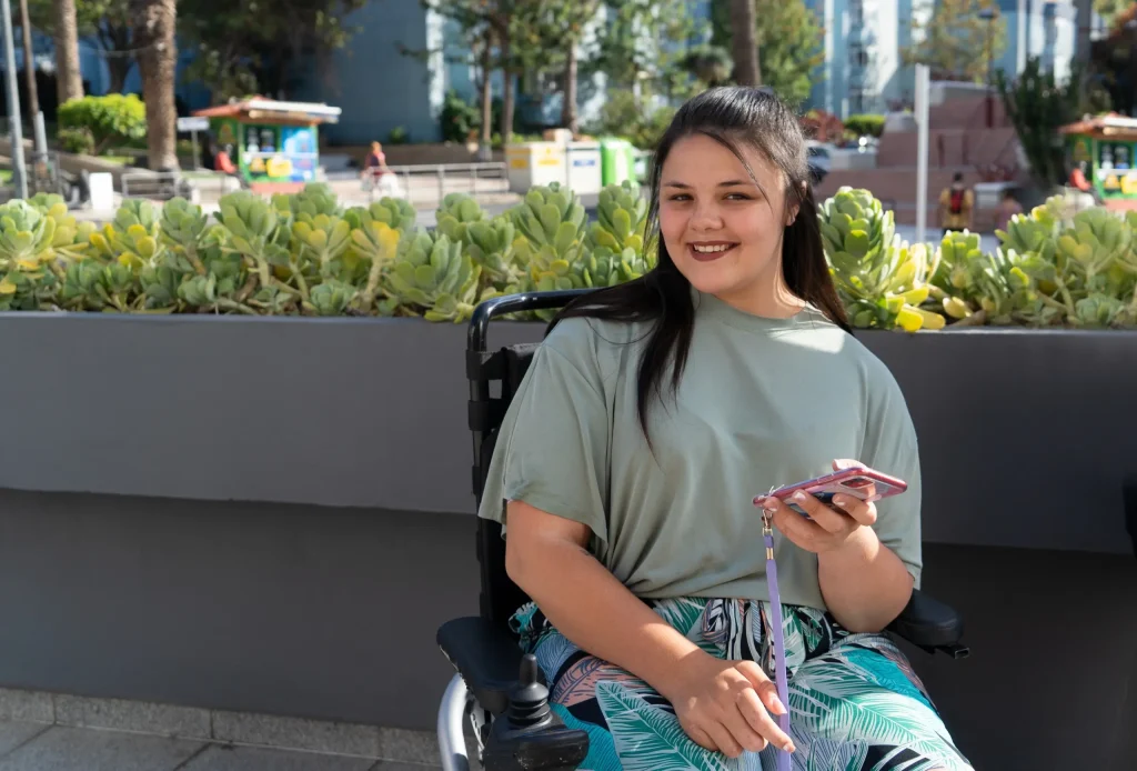 A woman in a wheelchair sits outdoors, holding a smartphone and smiling, with greenery and an urban background behind her—reflecting the quality of modern disability care services.